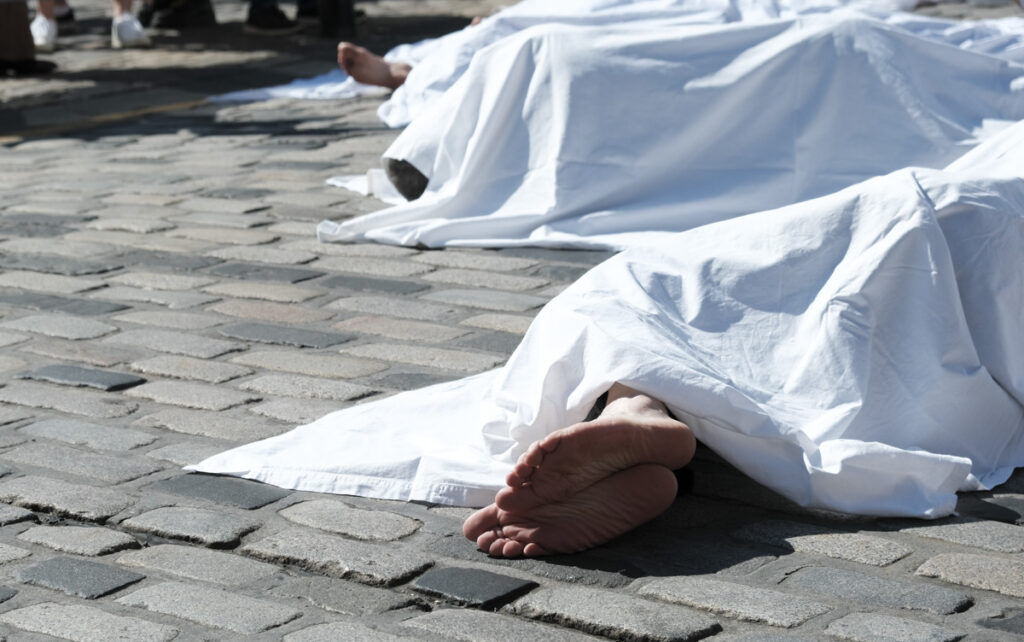 Close-up of XR Die-In participant lying on cobbles under a sheet, with bare feet visible