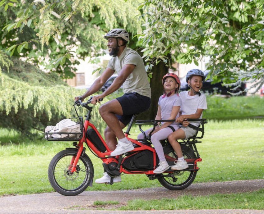 A man riding a Tern Longtail through a park with two girls riding on the back passenger seat.